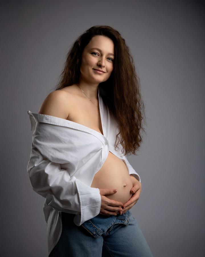Kobieta w ciąży z długimi brązowymi włosami w białej koszuli podczas sesji ciążowej.