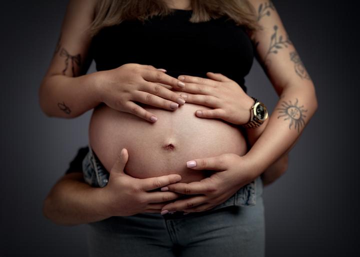 Kobieta w ciąży z tatuażami na ramionach delikatnie tuli brzuch podczas sesji ciążowej.