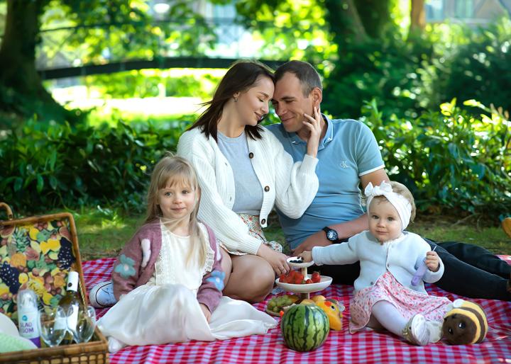 Rodzina na pikniku w zielonym ogrodzie, rodzice z dwójką małych dzieci przy kocu i owocami.