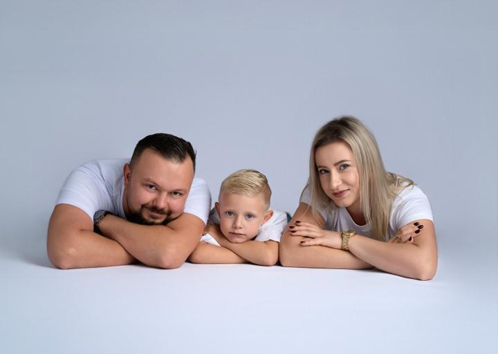 Rodzina z małym chłopcem w białych koszulkach pozuje przy błękitnym tle na sesji rodzinnej.