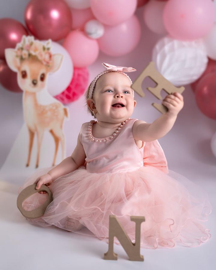 Laughing little girl in a pink tulle dress holding a wooden letter E, pink balloons behind her