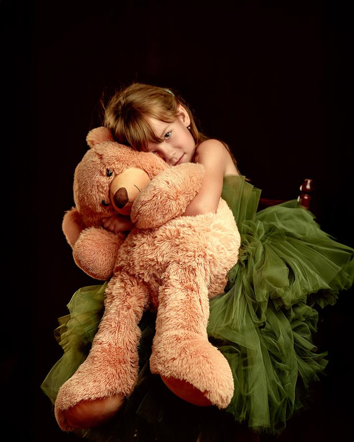 Girl in a green tulle dress hugging a large teddy bear, dark background