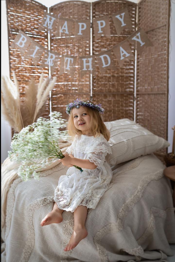 Girl in a white lace dress and flower crown sitting with a bouquet of baby's breath under a HAPPY BIRTHDAY banner
