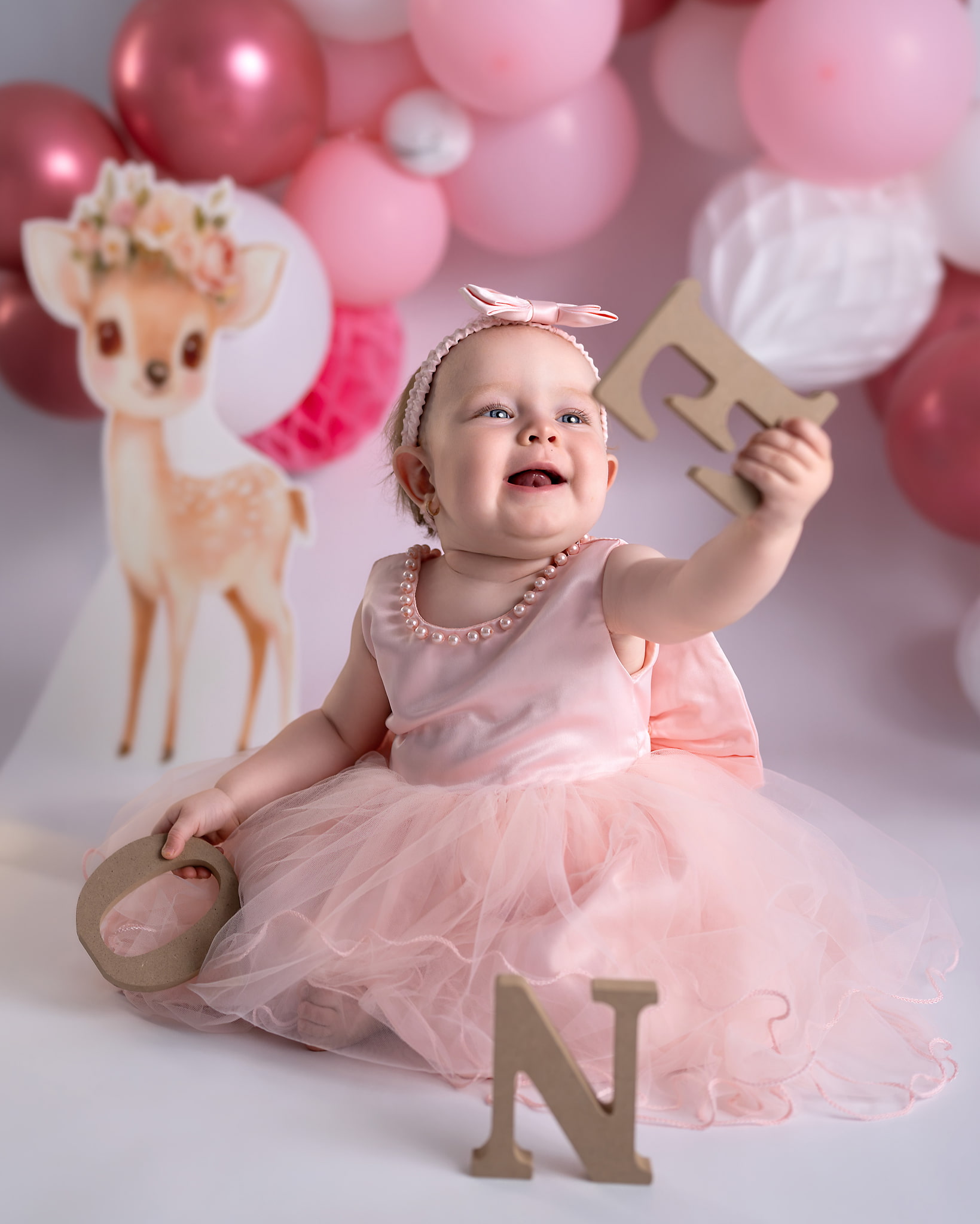 Lachend meisje in een roze tule jurkje met een houten letter E, roze ballonnen op de achtergrond