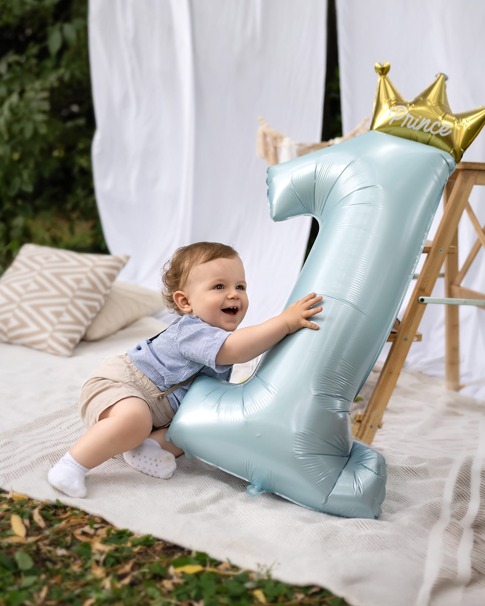 Smiling little boy hugging a blue number 1 balloon with a gold crown in a garden
