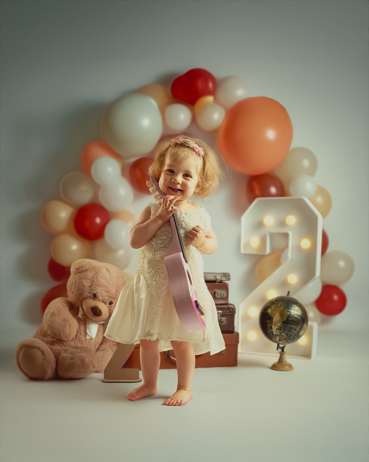 Meisje in een crèmekleurige jurk onder een ballonnenboog, naast een lichtgevend cijfer 2 en een teddybeer